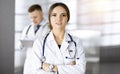 Portrait of a professional friendly woman doctor with a stethoscope, standing with crossed arms in a sunny clinic. Young