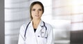 Portrait of a professional friendly woman doctor with a stethoscope, standing straight in a sunny clinic. Physician at