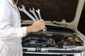 Portrait of professional young mechanic man in white uniform holding wrenches against car in open hood at the repair garage. Royalty Free Stock Photo