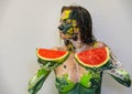 Portrait in profile of a sexy, yellow, green painted woman with large melons holding 2 fresh juicy red watermelon slices in her Royalty Free Stock Photo