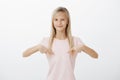 Portrait of proud fashionable girl with blond hair pointing at pink t-shirt with pleased and happy expression, showing Royalty Free Stock Photo