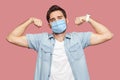 Portrait of proud satisfied young man with surgical medical mask in blue shirt standing in body building pose and showing his