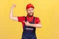 Strong worker man wearing blue uniform and red cap standing with raised arms and pointing at biceps.