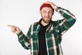 Portrait of puzzled guy wearing hat and plaid shirt laughing and pointing finger aside, while standing over white Royalty Free Stock Photo