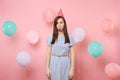 Portrait of puzzled upset young woman with party whistle in birthday hat and blue dress on pastel pink background with