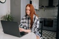 Portrait of puzzled young woman typing on laptop computer sitting at table, pensive looking to screen in kitchen Royalty Free Stock Photo