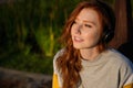 Portrait of a red-haired girl with freckles, green eyes in headphones, sitting in the sunset light and looking into the Royalty Free Stock Photo