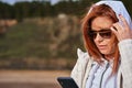 Portrait of a red-haired middle-aged woman in sunglasses and a raincoat looking at a smartphone. Sunny spring morning. Royalty Free Stock Photo