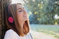 Portrait of red haired teenage girl listening to music in big pink earphones outdoors leaning to a tree Royalty Free Stock Photo