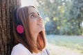 Portrait of red haired teenage girl listening to music in big pink earphones outdoors leaning to a tree Royalty Free Stock Photo