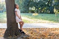 Portrait of red haired teenage girl in white short dress listening to music in big pink earphones outdoors leaning to a tree Royalty Free Stock Photo