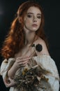 Portrait of a red-haired young woman in a vintage gold dress with bare shoulders with a dry bouquet in hands on a black background
