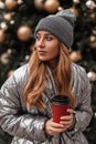 Portrait of a red-haired young woman in a vintage knitted gray hat in a fashionable silver jacket with a cup of coffee in hands