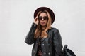Portrait of a redhaired sexy young hipster woman in an elegant hat in dark sunglasses in a trendy leather jacket with a backpack Royalty Free Stock Photo