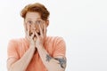 Portrait of redhead young man with freckles covering face with palms and peeking through fingers, screaming from fear