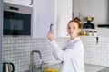 Portrait of redhead young woman opening door of kitchen cabinet at light kitchen room. Royalty Free Stock Photo