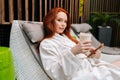 Portrait of redhead young woman wearing bathrobe relaxing with smartphone lying on lounger by poolside and drinking Royalty Free Stock Photo