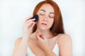 Portrait of redheaded young woman looking natural applying skin foundation with makeup sponge standing on white