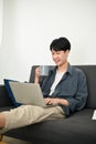 Portrait, Relaxed Asian man remote working from home, sipping coffee, using laptop on sofa Royalty Free Stock Photo