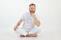 Relaxed sporty middle-aged bearded man with short grey hair sitting with crossed legs on floor on white background.