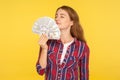 Portrait of rich greedy woman in casual checkered shirt holding money, smelling dollar banknotes with pleased expression