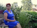 Portrait of 60s mature woman in blue dress sitting next to flower beds with daisies in village Royalty Free Stock Photo