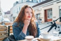 Portrait of sad red curled long hair caucasian teen girl sitting on a cozy cafe outdoor terrace on the street and looking at the Royalty Free Stock Photo