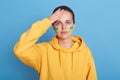 Portrait of sad woman in yellow hoodie with Ukrainian flag on cheeks, keeping her hand on head, worries about his country, posing