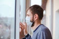 Sad young man in a medical mask who looks out the window through the window, he observes quarantine