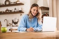 Portrait of satisfied blond woman 20s wearing casual sweatshirt working on laptop and writing down notes at home Royalty Free Stock Photo