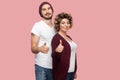 Portrait of satisfied couple of friends in casual style standing, hugging and showing thumbs up sign, looking at camera