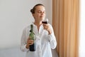 Portrait of satisfied delighted woman wearing white shirt holding bottle of alcoholic drink and smelling glass of wine, standing Royalty Free Stock Photo