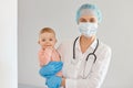 Portrait of satisfied doctor pediatrician wearing medical cap, surgical mask, gown and rubber gloves, standing with toddler girl