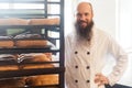 Portrait of satisfied young adult baker with long beard in white uniform standing in his workplace, near shelves with bread at the