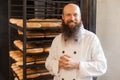 Portrait of satisfied young adult baker with long beard in white uniform standing in his workplace, near shelves with bread at the Royalty Free Stock Photo