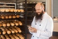 Portrait of satisfied young adult businessman baker with long beard in white uniform standing in bakery and have online order by