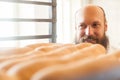 Portrait of satisfied young adult chef baker with long beard in white uniform standing in his bakery and looking on fresh bread in