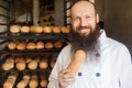 Portrait of satisfied young adult chef with long beard in white uniform standing in his bakery and holding fresh bread, looking at