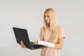 Portrait of scared blonde young woman with broken arm wrapped in plaster bandage holding laptop and working typing Royalty Free Stock Photo