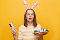 Portrait of scared shocked woman wearing bunny ears holding easter eggs and cell phone isolated over yellow background, using