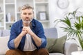 Portrait of a scary handsome gray-haired man in a blue shirt sitting on the sofa at home and seriously looking at the Royalty Free Stock Photo