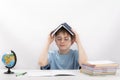 Portrait of schoolboy wearing glasses sits at the table and holding book over his head. Tired boy with pile of school books and