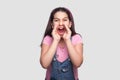 Portrait of screaming brunette young girl in casual style, pink t-shirt and blue denim overalls standing looking with hand near