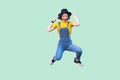 Portrait of screaming happy suprised young girl in blue denim overalls, yellow shirt and black hat jumping, looking and