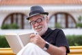 Portrait of a senior bearded man with hat sitting on a bench in the park relaxing and reading a book. Smiling carefree people Royalty Free Stock Photo