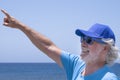 Portrait of a senior bearded man with white hair and blue cap and glasses smiling and standing at the beach pointing the finger at Royalty Free Stock Photo