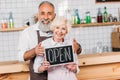 portrait of senior coffee shop owners in aprons holding open chalkboard together