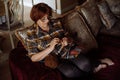 Portrait of senior concentrated woman with dark hair wearing checkered shirt, glasses, sitting on sofa, crocheting.