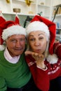 Portrait of senior couple in santa hat sitting on couch and blowing flying kisses at camera at home Royalty Free Stock Photo