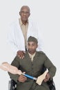 Portrait of senior doctor with US military officer holding artificial limb as he sits in wheelchair over gray background